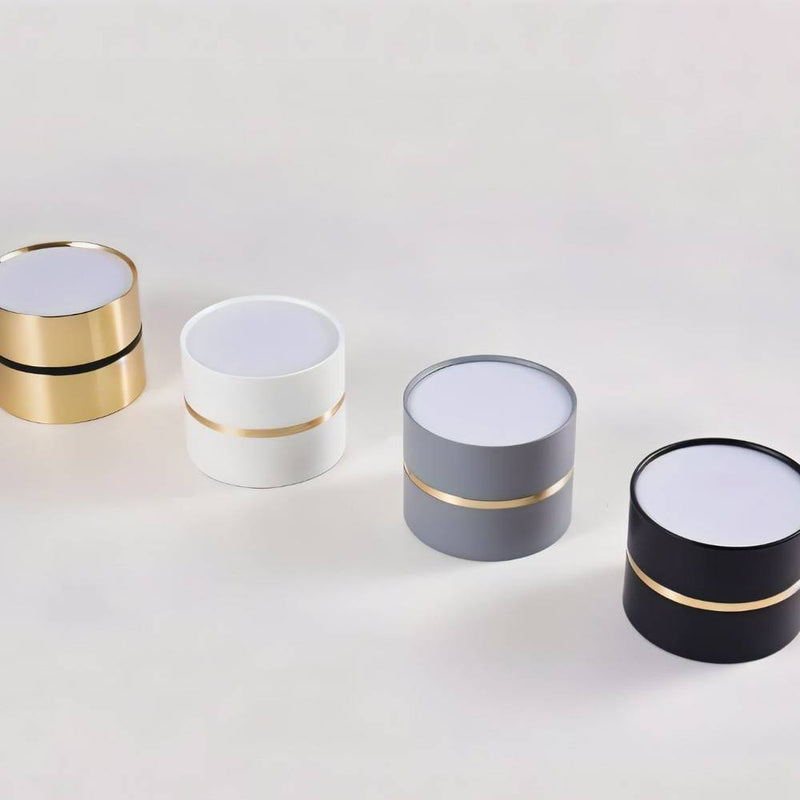 Four cylindrical downlights with white tops and gold accent bands in gold, white, gray, and black finishes on a neutral background.