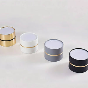Four cylindrical downlights with white tops and gold accent bands in gold, white, gray, and black finishes on a neutral background.