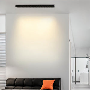 Linear black ceiling downlight illuminating a minimalist modern living room with white walls and dark sofa.