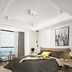 White square downlight fixtures mounted on a modern bedroom ceiling above a bed with gray and yellow bedding.