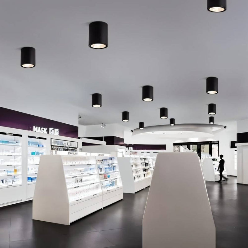 Black cylindrical ceiling downlights illuminating a modern pharmacy with white display cases and dark flooring.