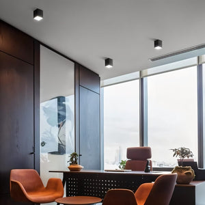 Black square downlight fixture mounted on gray ceiling in modern office with orange chairs and city view.