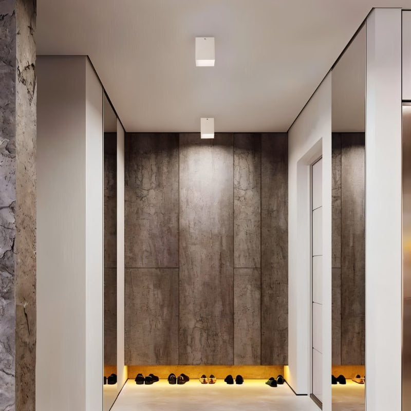 White square ceiling downlight mounted in a modern hallway with concrete walls and warm floor lighting.