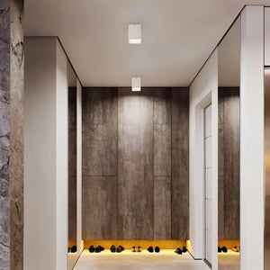 White square ceiling downlight mounted in a modern hallway with concrete walls and warm floor lighting.