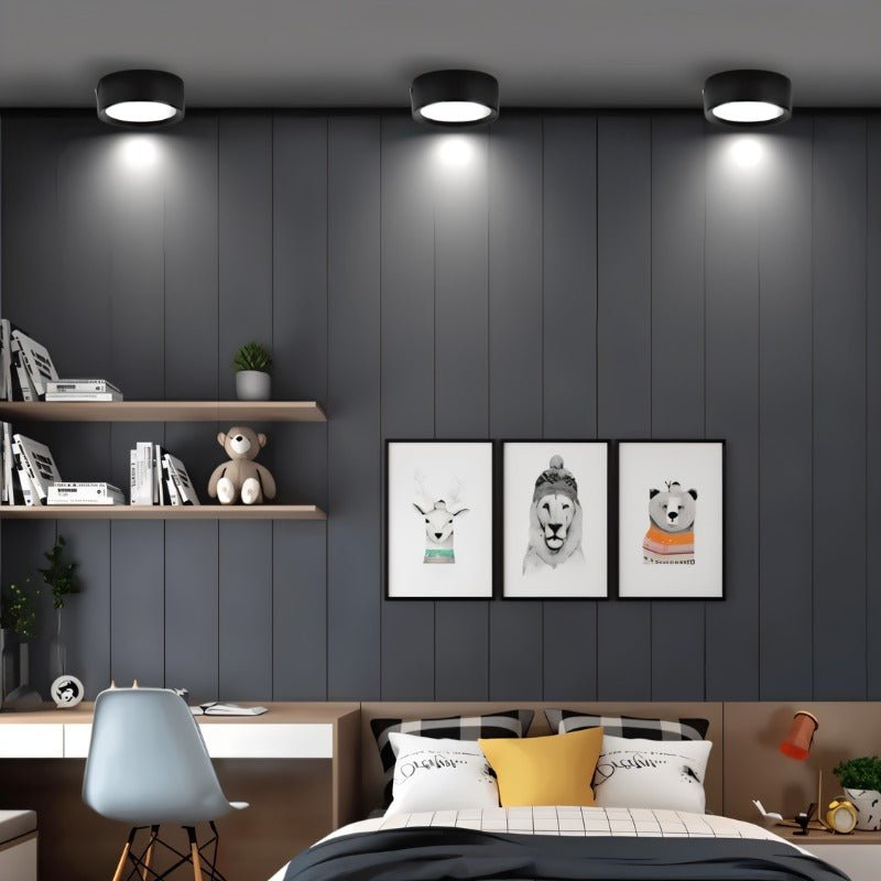 Black round ceiling downlight with white LED glow mounted above modern bedroom with gray walls and floating shelves.