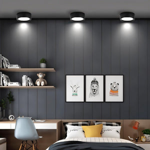 Black round ceiling downlight with white LED glow mounted above modern bedroom with gray walls and floating shelves.
